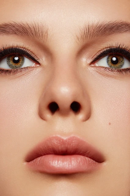 Close-up portrait of a woman's face featuring hazel eyes, light pink lips, and fair skin with natural makeup in a soft, diffused light.