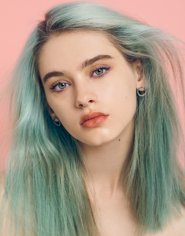Close-up portrait of a young adult woman with long, light blue hair and fair skin against a soft pink background in a beauty context.