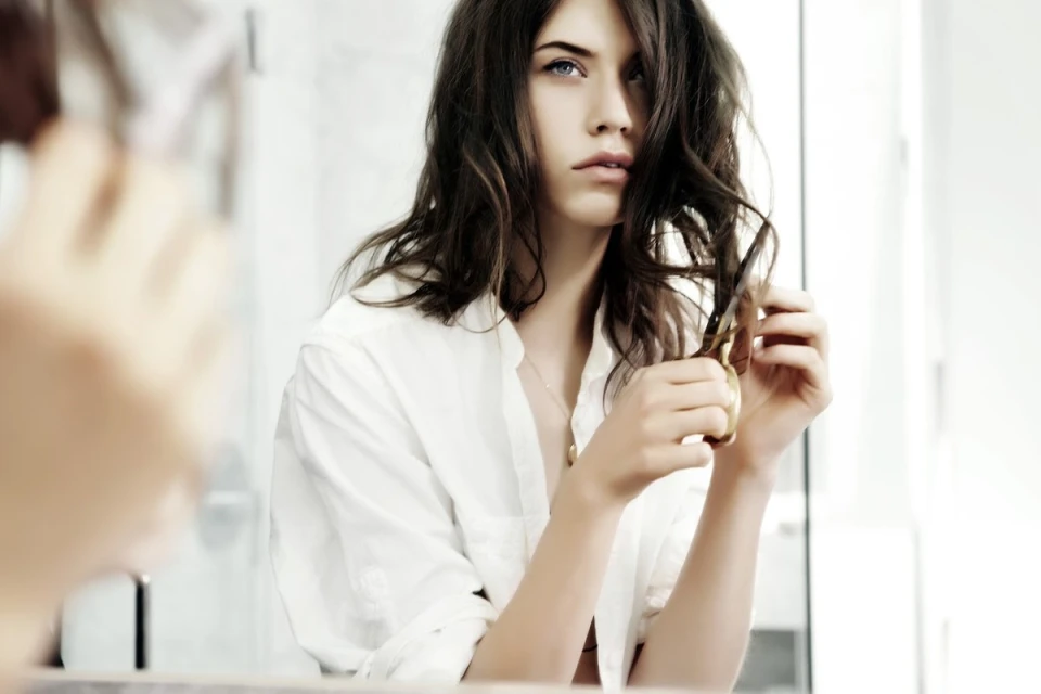 Young woman with wavy brown hair wearing a white shirt cuts a strand of hair with gold scissors indoors with light, airy background.
