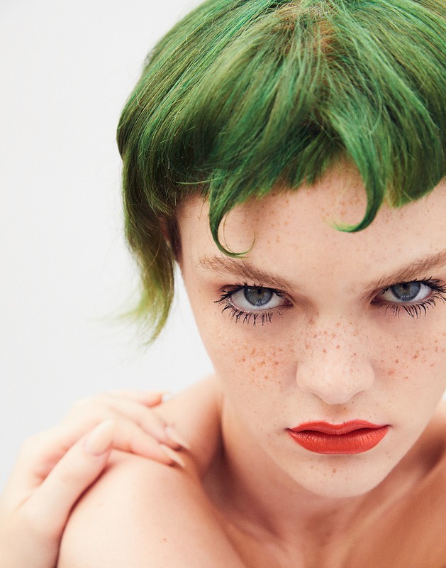 Close-up portrait of a young adult woman with short, green hair and freckles, looking directly at the camera against a light grey backdrop.