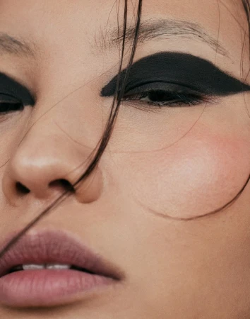 Close-up beauty portrait of a woman featuring heavy black graphic eyeliner and subtle pink blush, with loose strands of hair across her face.