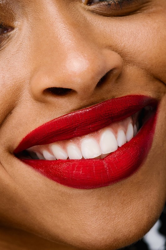 Close-up of an adult female smiling widely, showcasing her white teeth and bright red lipstick, captured in soft, flattering lighting.