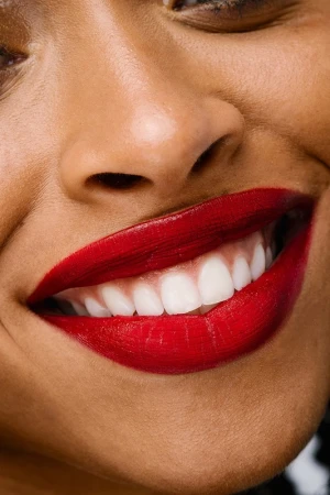 Close-up of an adult female smiling widely, showcasing her white teeth and bright red lipstick, captured in soft, flattering lighting.