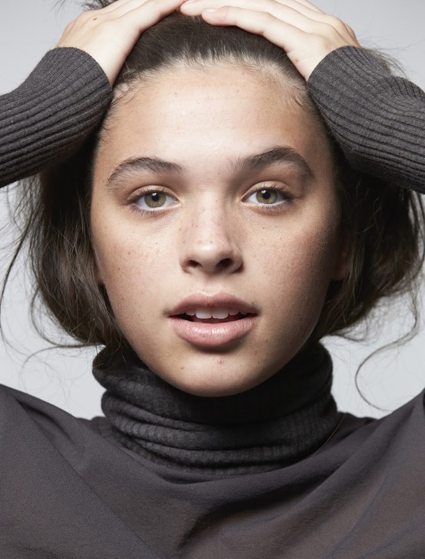 Close-up portrait of a young adult woman with hands on her head, wearing a high-neck top, against a neutral studio backdrop.