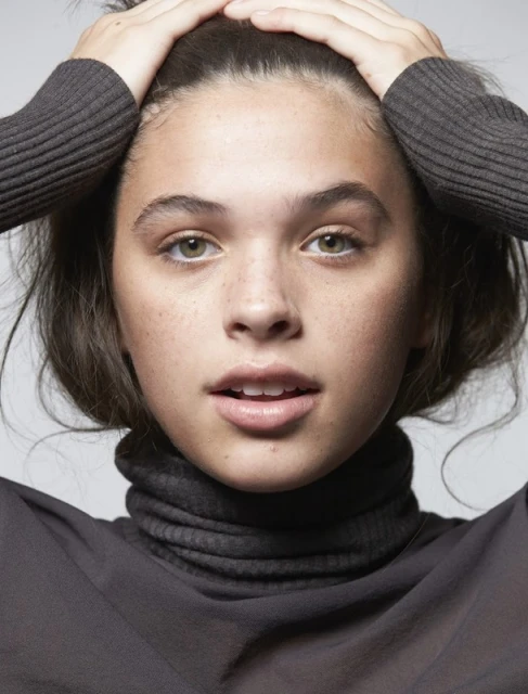 Close-up portrait of a young adult woman with hands on her head, wearing a high-neck top, against a neutral studio backdrop.