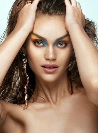 Close-up portrait of a woman with vibrant blue and yellow eye makeup holding wet hair against a pale background