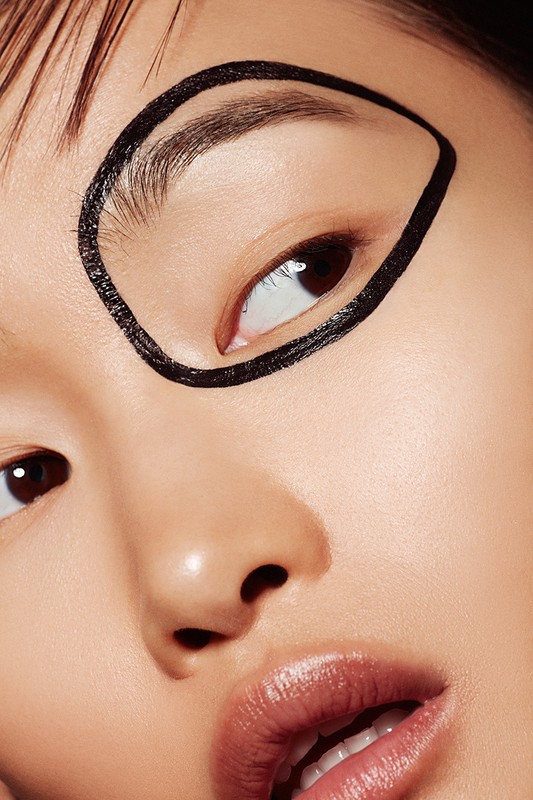 Close-up portrait of a young adult woman with black eyeliner around her eye, glossy lips, and visible makeup. Beauty and fashion editorial.