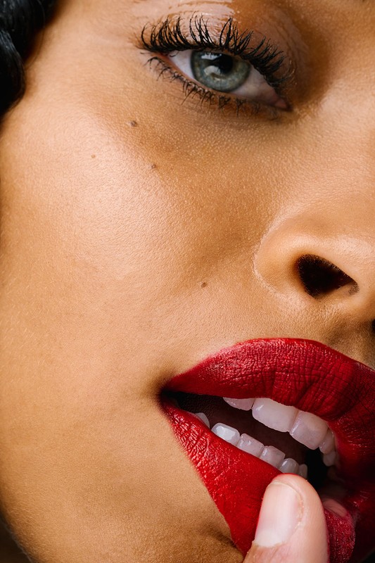 Extreme close-up of a woman with fair skin and gray eyes, featuring red lipstick and a finger gently touching her lower lip.