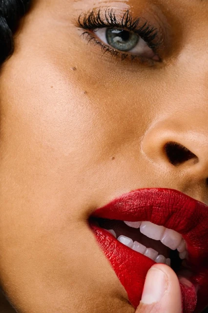 Extreme close-up of a woman with fair skin and gray eyes, featuring red lipstick and a finger gently touching her lower lip.