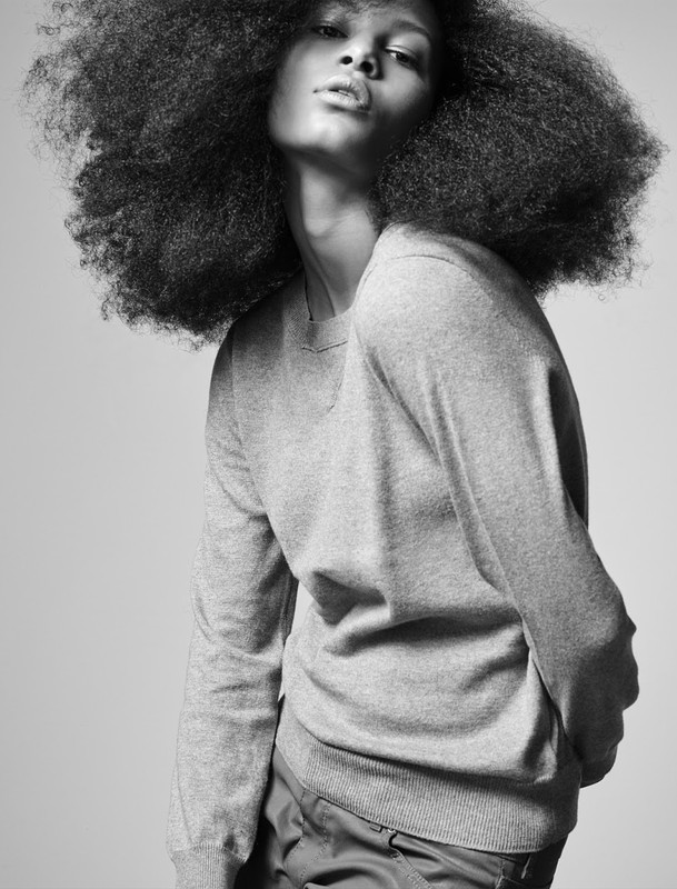 Black and white portrait of a person with an Afro hairstyle wearing a grey sweater and dark pants against a light grey background.