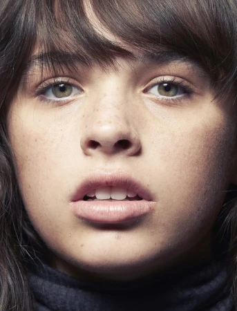 Close-up portrait of a young woman with dark hair, bangs, green eyes, and light freckles against a neutral background.