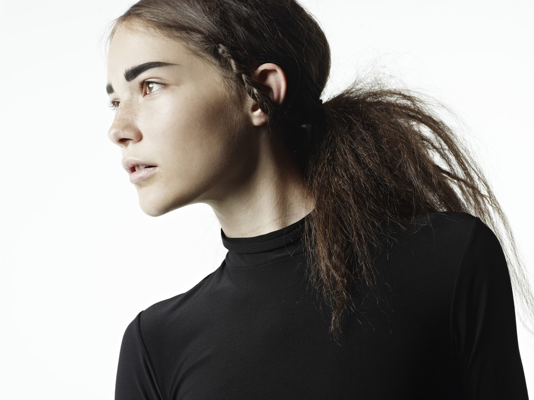 Profile portrait of a young female model with dark hair in a ponytail and bold brows wearing a black top against white background.