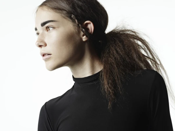 Profile portrait of a young female model with dark hair in a ponytail and bold brows wearing a black top against white background.