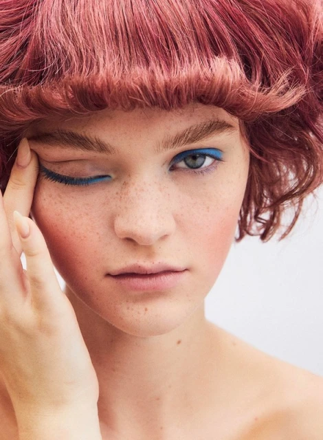 Close-up of a woman with red hair and blue eyeliner, covering one eye with her fingers, against a plain white background.
