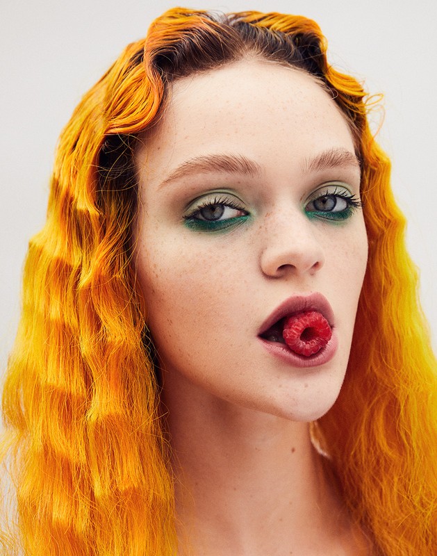 A close-up portrait of a young woman with dyed orange wavy hair, green eyeshadow, and a raspberry in her mouth against a white backdrop.