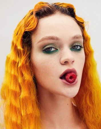 A close-up portrait of a young woman with dyed orange wavy hair, green eyeshadow, and a raspberry in her mouth against a white backdrop.