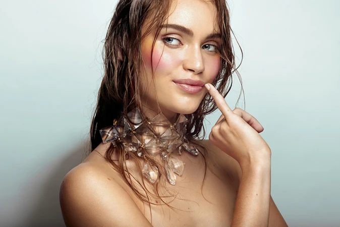Close-up portrait of a woman with wet hair, colorful cheek makeup, and a clear geometric crystal choker necklace