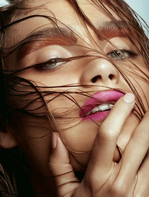 Close-up portrait of a model with wet brown hair partially covering her face, pink lipstick, and bronze eye makeup in a beauty style.