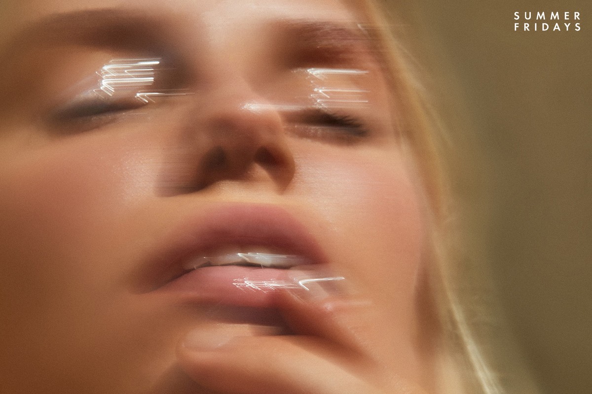 Close-up portrait of a blonde woman with eyes closed, glossy lips, and a finger near her mouth in soft, neutral light.