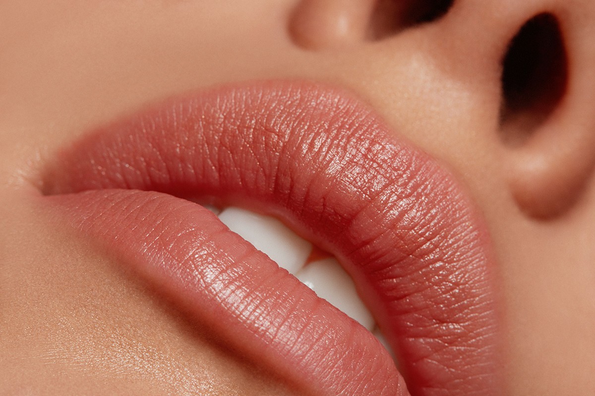 Close-up of a woman's face featuring glossy pink lips, white teeth, smooth skin texture, and defined nostrils, with a focus on beauty and skincare.