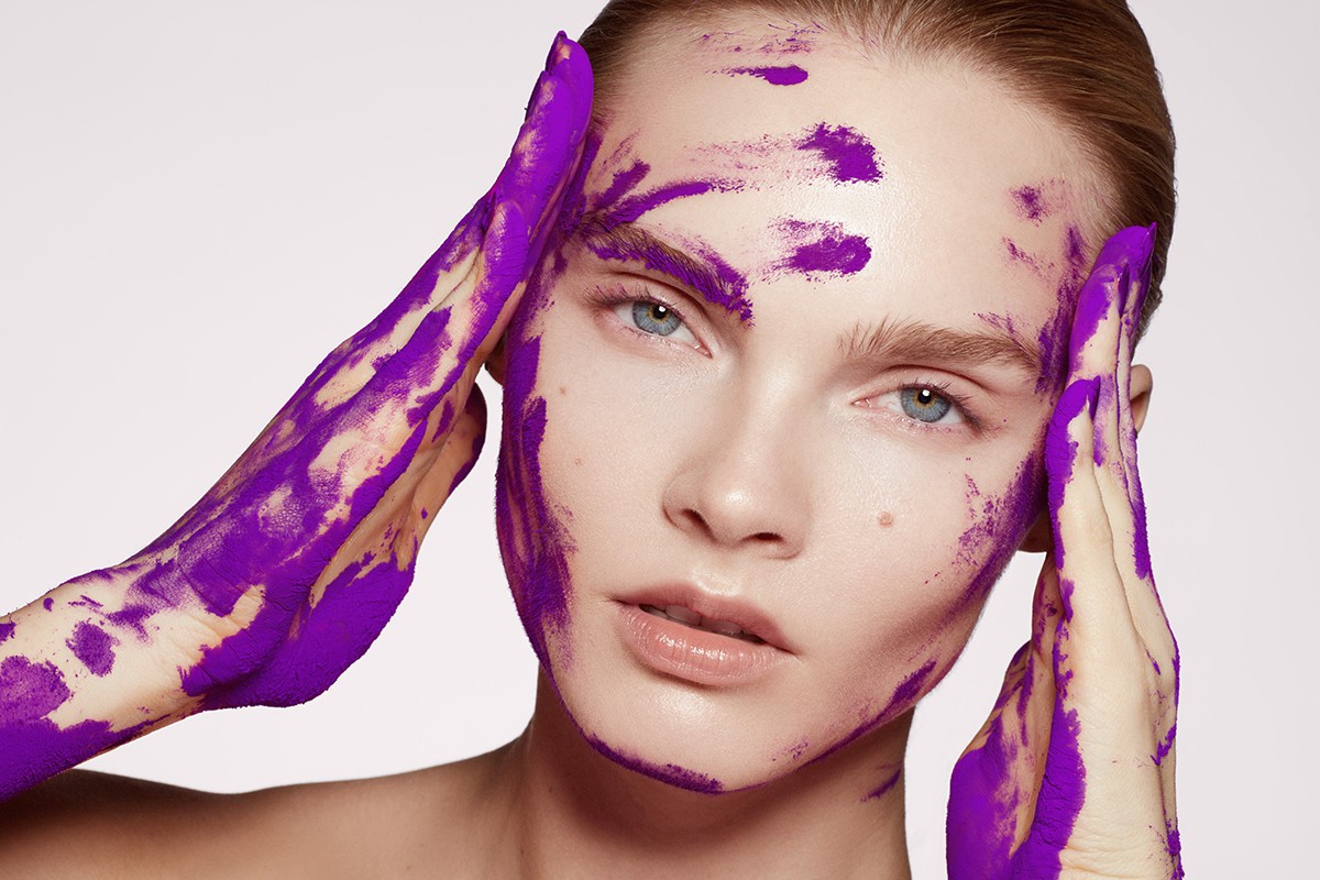 Close-up beauty shot of a woman with purple paint smeared on her face and hands in a photo studio with soft, diffused lighting.