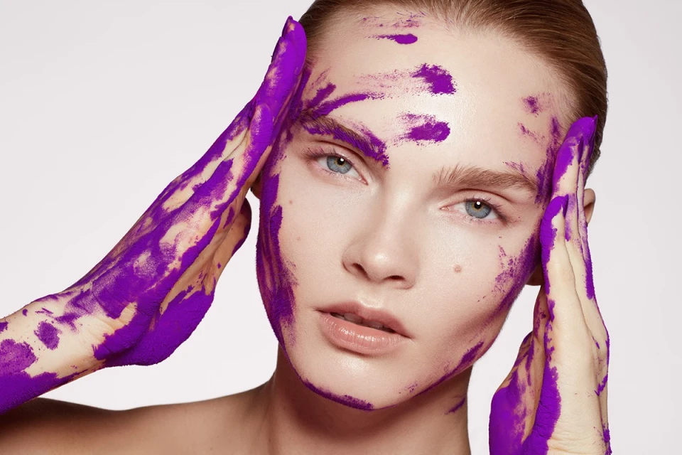 Close-up beauty shot of a woman with purple paint smeared on her face and hands in a photo studio with soft, diffused lighting.