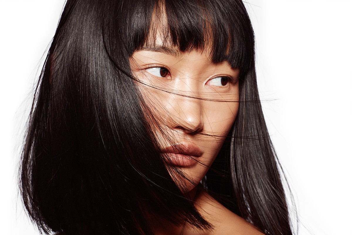 Close-up portrait of a young Asian woman with dark, straight hair and bangs, looking off to the side against a white background.