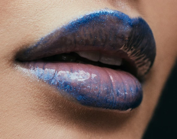 Close-up portrait of lips wearing glossy metallic blue lipstick with visible glitter and teeth showing slightly.