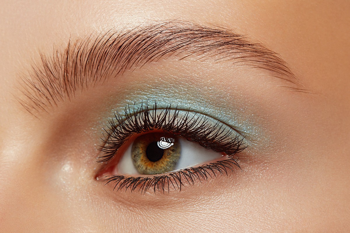 Close-up shot of a woman's eye with green-hazel iris, long dark lashes, expertly shaped eyebrow, and light-blue eye shadow.
