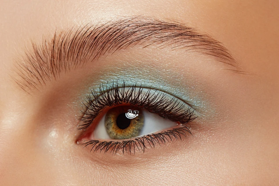 Close-up shot of a woman's eye with green-hazel iris, long dark lashes, expertly shaped eyebrow, and light-blue eye shadow.