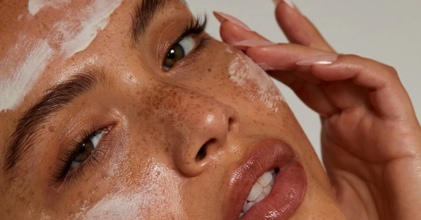 Close-up of a woman's face with skincare product application, featuring freckles, glossy lips, manicured hand, and neutral background.