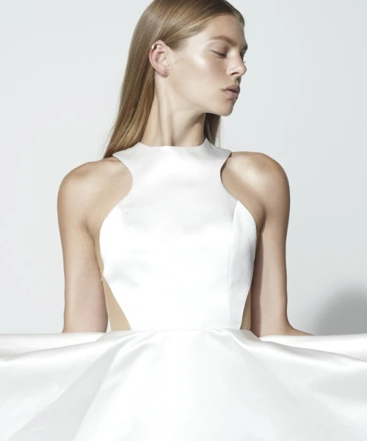 Blonde woman in a white dress with high neck poses in a studio with a white background. The dress features a fitted bodice and full skirt.