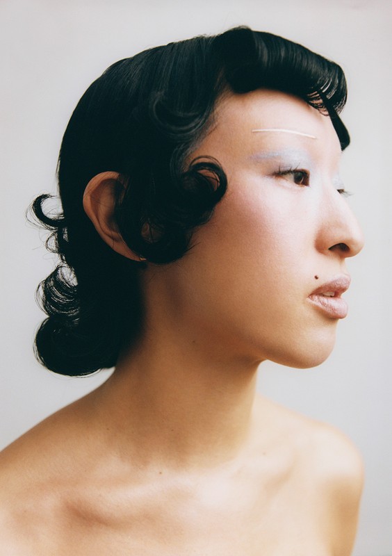 Portrait of an Asian woman with black wavy hair and striking white makeup on her eyebrows and eyelids, looking to the side against a plain background.