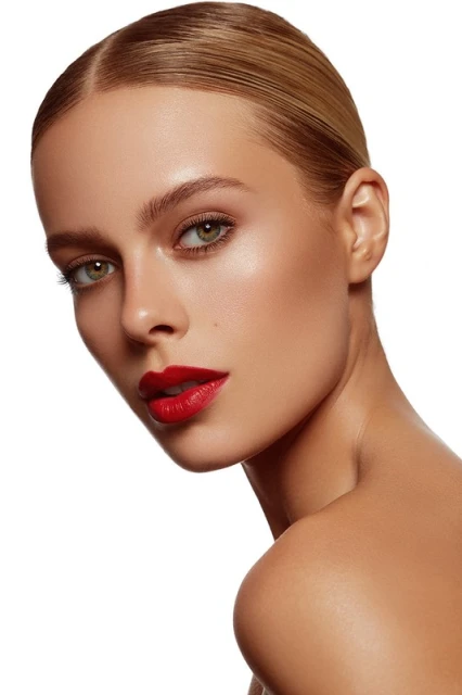 Close-up portrait of a young adult woman with sleek blonde hair, red lipstick, and flawless makeup against a bright white backdrop.