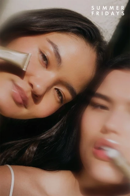 Close-up of two adult females with dark hair, one holding a cosmetic product; soft lighting and shallow depth of field enhance the beauty.