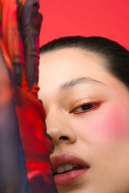 Close-up of a woman's face with red eyeliner and blush, and a textured, semi-transparent fabric partially covering her face, vivid red backdrop.