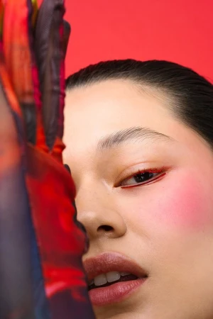 Close-up of a woman's face with red eyeliner and blush, and a textured, semi-transparent fabric partially covering her face, vivid red backdrop.