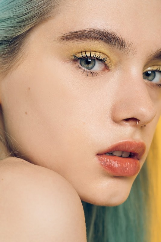 Close-up portrait of a young adult female with yellow eye makeup, a nose ring, and light teal hair, against a gradient yellow background.