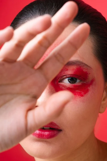 Close-up of a woman's face with bright red makeup around her eyes and red lipstick, partially obscured by her outstretched hand against a red background.