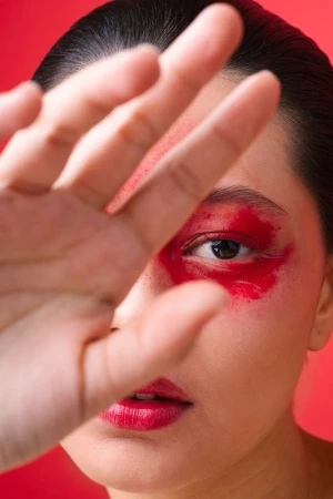 Close-up of a woman's face with bright red makeup around her eyes and red lipstick, partially obscured by her outstretched hand against a red background.