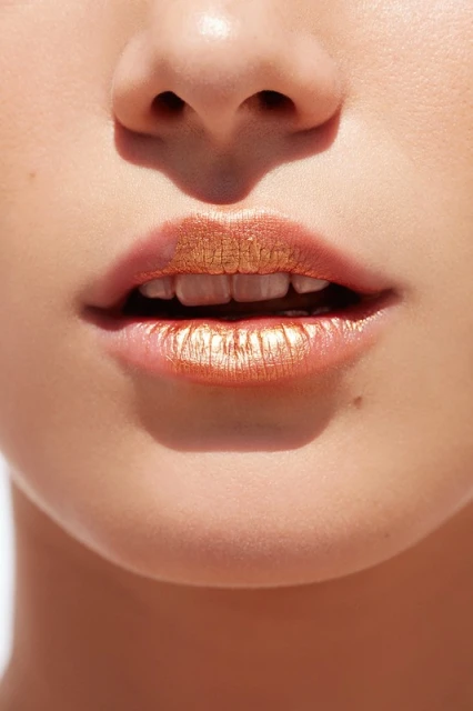 Close-up shot of a woman's face showing her lips with shiny, gold lipstick, a small portion of her nose, and fair skin.