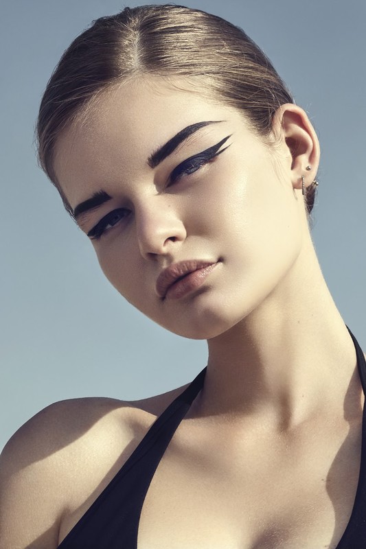Close-up portrait of a young adult female with dramatic winged eyeliner and neutral makeup against a plain blue background.