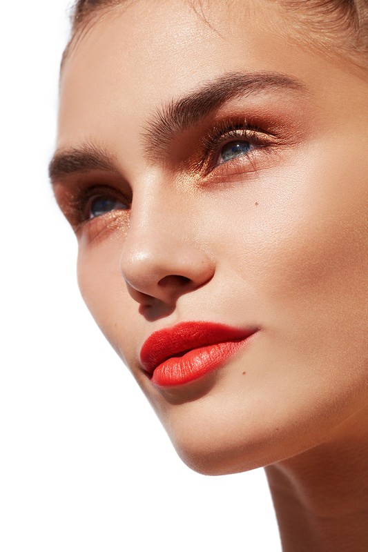 Close-up portrait of a young adult female with bright red lipstick and gold eyeshadow makeup, looking upward against a white background.