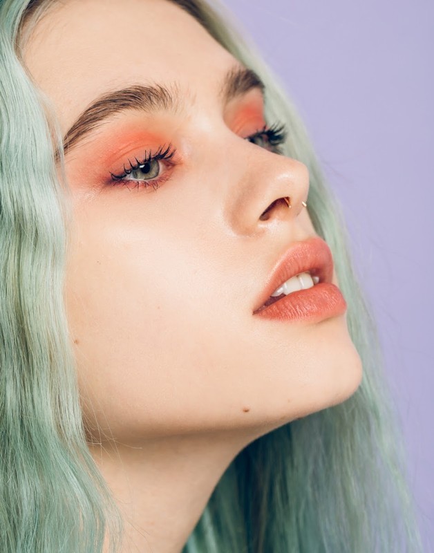 Close-up of a young woman with green-blue wavy hair, coral-toned eye makeup, and a piercing on her nose, posing in a beauty shot.