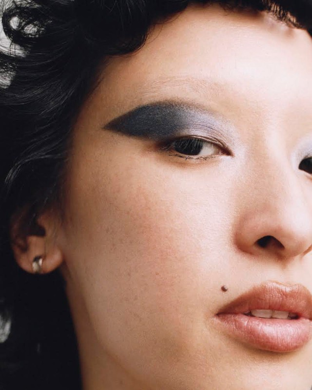 Close-up portrait of a woman with dramatic dark winged eye makeup and a small beauty mark near her lip.