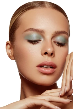 Close-up of a young woman with light skin and blue eyeshadow, gazing downward with her hands near her face on a white background.
