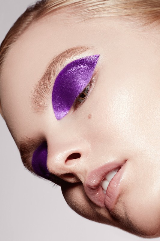 Close-up of a woman's face with artistic purple eyeshadow covering the eyelid and under-eye area, set against a plain background.