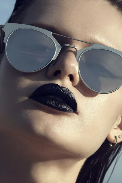 Close-up of an adult woman wearing sunglasses and dark lipstick, highlighting fashion and beauty elements against a bright sky.