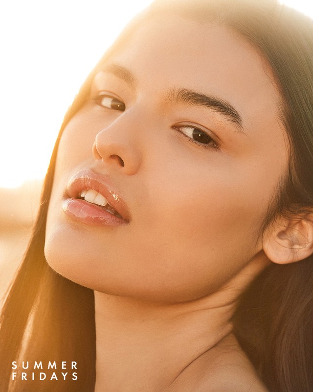 Close-up portrait of a young woman with glowing skin and glossy lips against a sunlit backdrop, showcasing beauty and summer vibes.