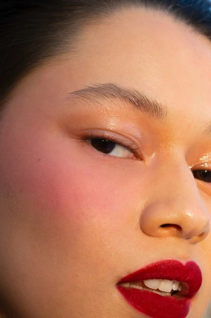 Close-up studio shot of a woman with red lipstick and pink blush. The woman has one eye open and fair skin with a glossy look.